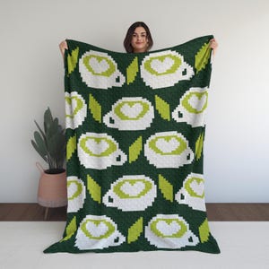 May include: A dark green blanket featuring a repeating pattern of white pixelated coffee cups with green heart-shaped latte art and green leaves. The blanket is held up by a person.