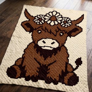 May include: A rectangular cream-colored blanket featuring a pixel art design of a brown highland cow. The cow has a white muzzle and a crown of white daisies between its horns. The blanket has a textured, dimpled surface throughout the fabric.
