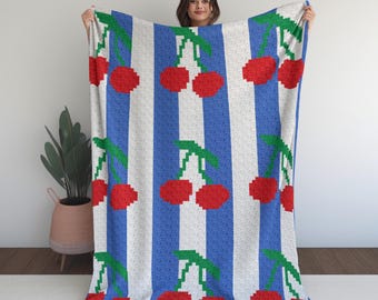 May include: A soft blanket with a repeating pattern of red cherries on green stems against a blue and white striped background. The blanket is held up, showcasing the design and texture.