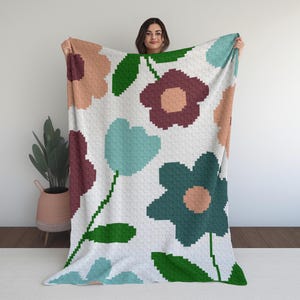 May include: A white blanket with a repeating floral pattern in shades of teal, burgundy, peach, and green. The flowers have a pixelated design, and the blanket is held up by a person.