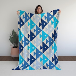 May include: A large, soft blanket with a geometric pattern in shades of blue and off-white. The design features repeating shapes resembling fish or arrows, creating a textured appearance. The blanket is held up, showcasing its size and the intricate pattern.