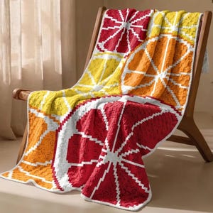 Op de afbeelding: Een kleurrijke gehaakte deken met pixelachtige citrusvruchten in rood, oranje en geel, met witte accenten. De deken ligt over een houten stoel en is een gezellig woonaccessoire.