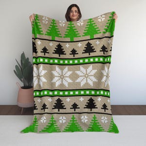 May include: A beige blanket with a green and black Christmas tree pattern, white snowflakes, and green and black stripes. The blanket is held up by a person.