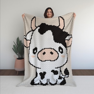 May include: A beige blanket with a large, pixelated cartoon cow design. The cow has a black and white pattern with pink accents on its ears and cheeks. The blanket is held up, displaying the full design.