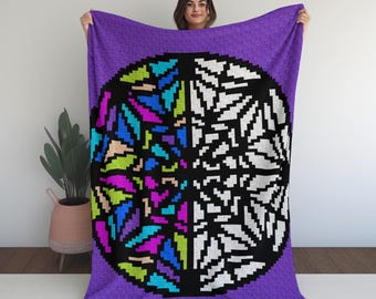 May include: A purple throw blanket featuring a pixelated design of a circle divided in half. One side has colorful geometric shapes, the other side is white. The blanket is held up by a person.