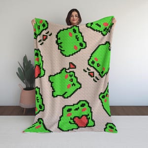 May include: A beige blanket featuring a repeating pattern of pixelated green frogs with red hearts. The frogs have black outlines and are in various poses. The blanket is held up, showcasing the design.