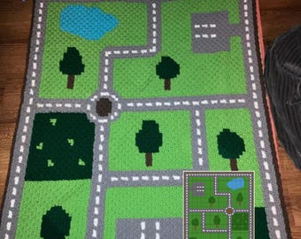 May include: A green crocheted play mat with a city design featuring roads, trees, and a pond. The mat has a gray border and white lines for roads. The text "Customer Photo" is at the bottom left corner of the mat.