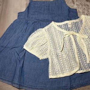 May include: A light blue denim dress with a white crocheted cardigan. The cardigan has a bow at the neckline and short sleeves.