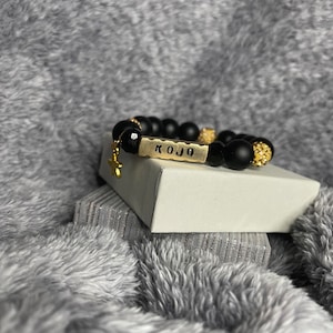 Könnte beinhalten: Schwarzes Perlenarmband mit einem goldenen Anhänger und einem goldenen Namensschild mit der Aufschrift "KOJO".