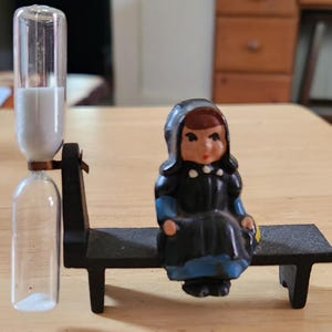 May include: A clear hourglass with white sand and a black metal stand next to a small figurine of a person in a black dress and blue accents sitting on a black bench. The figurine has brown hair and a white bonnet.