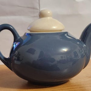 May include: A blue ceramic teapot with a cream-colored lid and knob. The teapot has a rounded body, a curved handle, and a spout. The glossy surface reflects the surrounding environment. A classic design for serving tea.