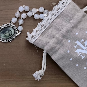 May include: A white rosary with a silver oval pendant featuring a black and white photo of a baby. The rosary is in a small, beige drawstring bag with white lace trim and the text "JMJ" and a heart.