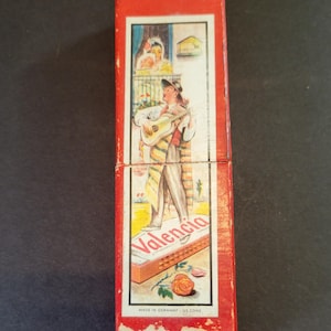 May include: Vintage red box with a colorful illustration of a man playing a guitar in front of a balcony. The word "Valencia" is printed on the box, along with the text "Made in Germany - US Zone".