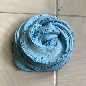 May include: A close-up of a blue slime with small blue beads embedded in it. The slime is on a white tiled surface.