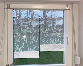May include: White window curtains with a floral pattern and a decorative border. The curtains are hanging in front of a window with a view of trees and a grassy area. The curtains are made of a sheer fabric.