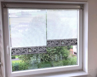 May include: A white window with two light-colored roller shades. The shades have a decorative border with a dark gray and silver floral design. The window overlooks a green, tree-filled landscape.