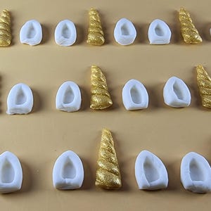 Puede incluir: Filas de conjuntos de cuernos y orejas de unicornio. Los cuernos son dorados con un diseño en espiral texturizado. Las orejas son blancas, de forma ovalada y tienen un centro hueco. Los conjuntos están dispuestos sobre una superficie marrón claro.