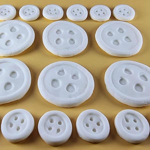 Può includere: Un assortimento di bottoni bianchi e rotondi con quattro fori ciascuno, disposti su una superficie marrone chiaro. I bottoni variano di dimensioni, con quelli più grandi al centro e quelli più piccoli in alto e in basso. I bottoni sono distanziati uniformemente.