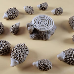 May include: A display of small, textured, grey hedgehog ornaments with white snouts, positioned around a miniature tree stump. The ornaments have a detailed, spiky appearance, and the stump has concentric rings.