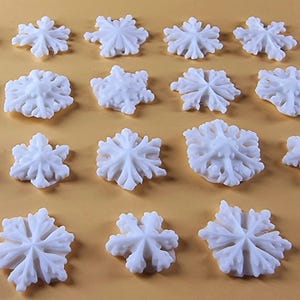 Peut inclure: Une collection de vingt-et-un flocons de neige blancs de tailles variées. Chaque flocon de neige présente un design détaillé en trois dimensions avec de multiples pointes et des motifs complexes. Les flocons de neige sont disposés sur une surface orange clair.