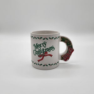 Puede incluir: Taza de cer&aacute;mica blanca con un borde de acebo verde y bayas rojas. La taza tiene una inscripci&oacute;n verde "Merry Christmas" con un lazo rojo. El asa tiene forma de corona navide&ntilde;a verde y roja.