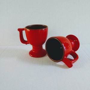 Op de afbeelding: Twee rode keramische bekers met een voet. De bekers zijn leeg en staan op een wit oppervlak.