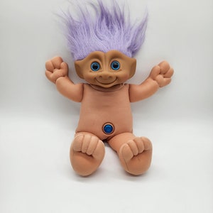 May include: A purple-haired troll doll with blue eyes and a light brown body. The doll is sitting with its legs crossed and its arms outstretched.
