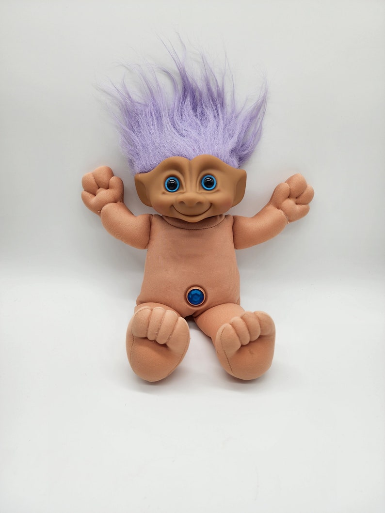 May include: A purple-haired troll doll with blue eyes and a light brown body. The doll is sitting with its legs crossed and its arms outstretched.