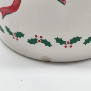 Puede incluir: Taza de cer&aacute;mica blanca con una cinta roja y hojas de acebo verdes con bayas rojas alrededor del borde.