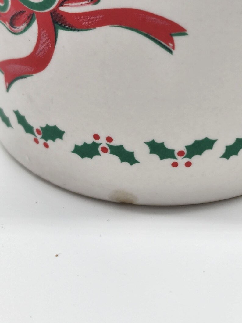 Puede incluir: Taza de cer&aacute;mica blanca con una cinta roja y hojas de acebo verdes con bayas rojas alrededor del borde.
