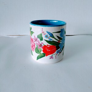 May include: A white ceramic mug with a blue rim and a floral design featuring pink, red, and blue flowers.