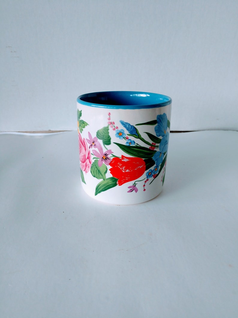 May include: A white ceramic mug with a blue rim and a floral design featuring pink, red, and blue flowers.