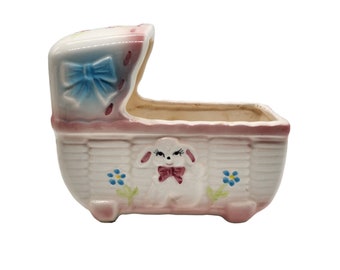 May include: A white ceramic baby crib planter with a pink and blue trim. The crib has a white lamb with a pink bow inside. The planter has a blue bow on the top.