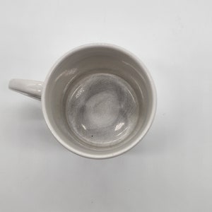 Puede incluir: Una taza de cer&aacute;mica blanca con asa. El interior de la taza est&aacute; vac&iacute;o y tiene un color gris claro.