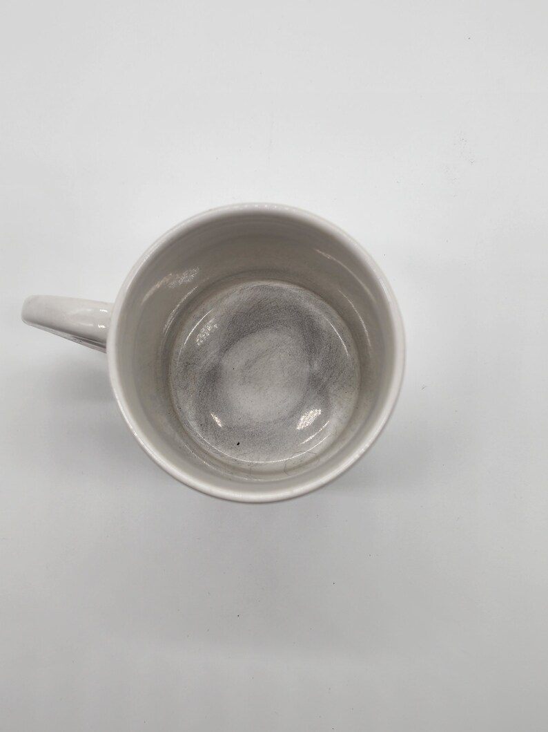 Puede incluir: Una taza de cer&aacute;mica blanca con asa. El interior de la taza est&aacute; vac&iacute;o y tiene un color gris claro.