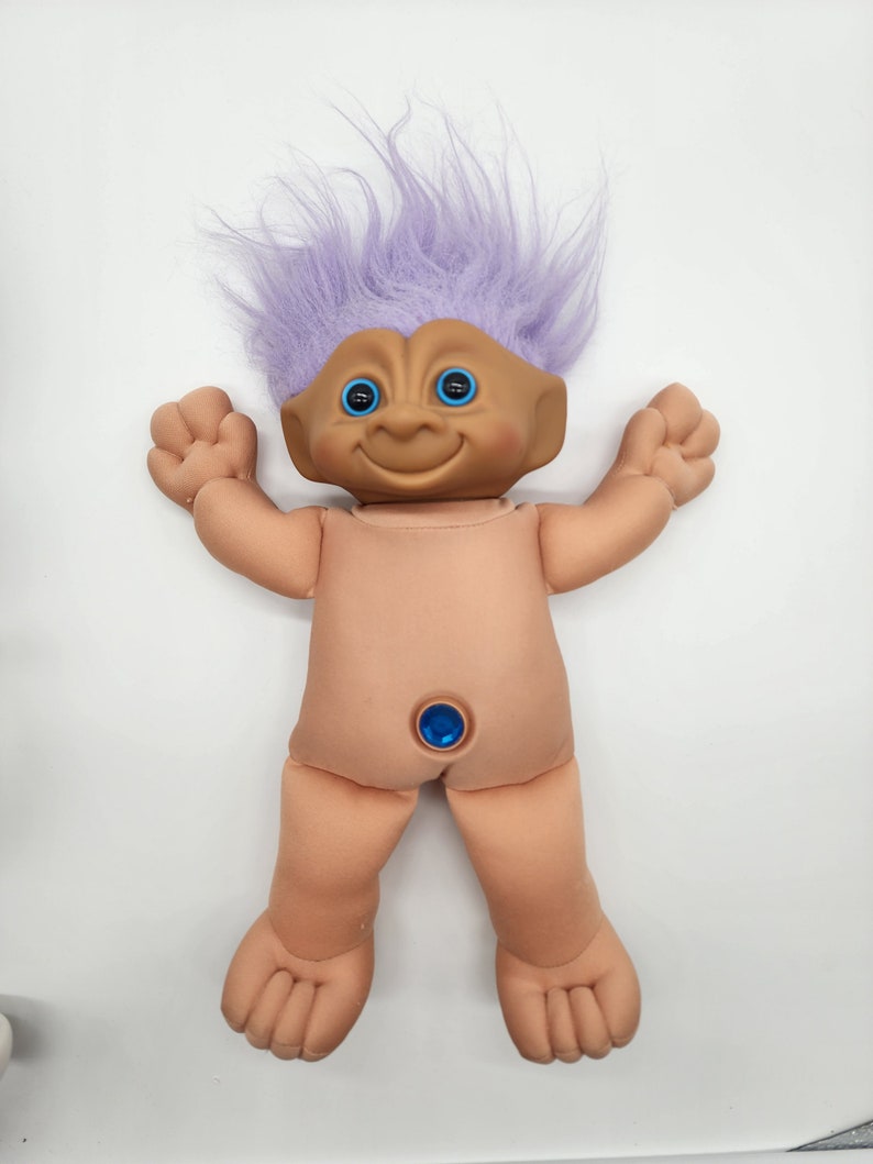 May include: A plush troll doll with purple hair and blue eyes. The doll has a blue gem on its belly button.