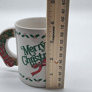 Puede incluir: Taza de cer&aacute;mica blanca con un dise&ntilde;o de acebo rojo y verde y las palabras "Merry Christmas" en letras verdes. La taza tiene un asa con forma de corona navide&ntilde;a roja y verde.