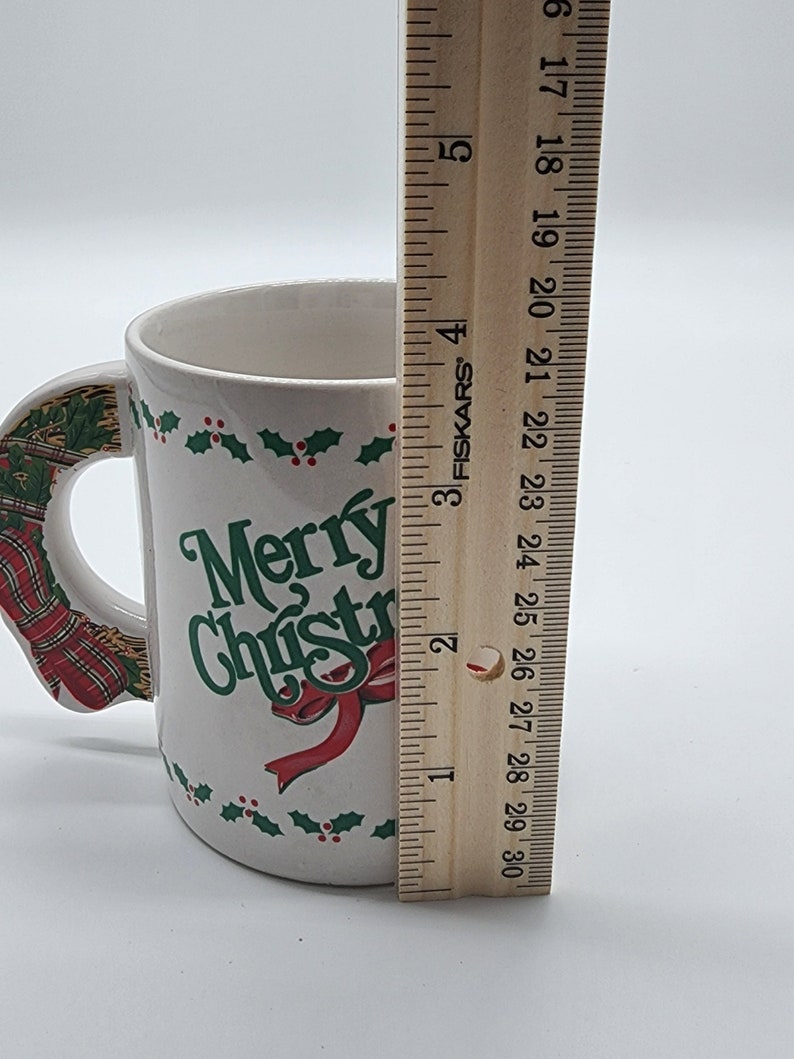 Puede incluir: Taza de cer&aacute;mica blanca con un dise&ntilde;o de acebo rojo y verde y las palabras "Merry Christmas" en letras verdes. La taza tiene un asa con forma de corona navide&ntilde;a roja y verde.