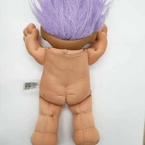 May include: A plush troll doll with purple hair. The doll is standing on a white surface.