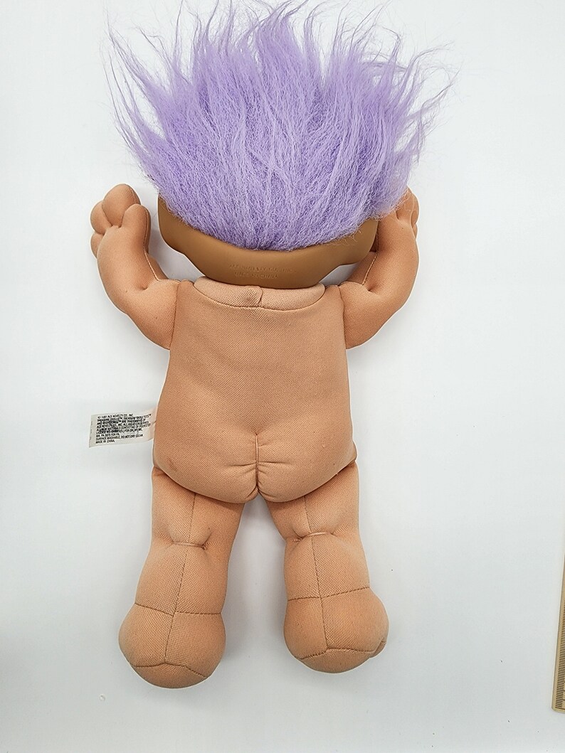 May include: A plush troll doll with purple hair. The doll is standing on a white surface.