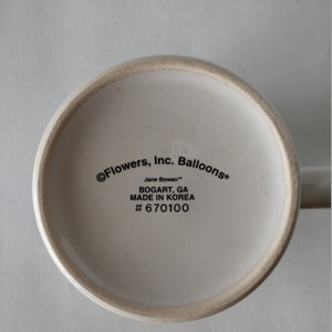 May include: White ceramic mug with black text on the bottom reading "&copy; Flowers, Inc. Balloons&reg; Jane Bowen&trade; BOGART, GA MADE IN KOREA # 670100"