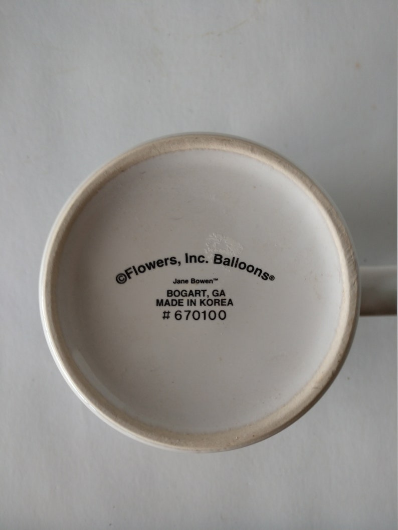 May include: White ceramic mug with black text on the bottom reading "&copy; Flowers, Inc. Balloons&reg; Jane Bowen&trade; BOGART, GA MADE IN KOREA # 670100"