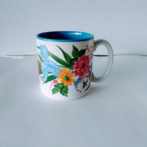 May include: A white ceramic mug with a blue rim and a floral design featuring pink, yellow, blue, and green flowers.