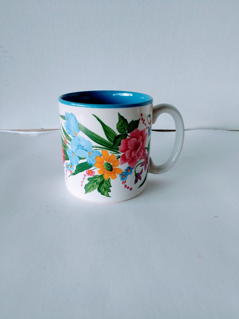 May include: A white ceramic mug with a blue rim and a floral design featuring pink, yellow, blue, and green flowers.
