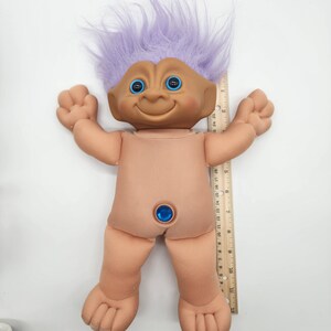 May include: A purple haired troll doll with blue eyes and a blue gem on its belly. The doll is about 12 inches tall and is standing next to a ruler.