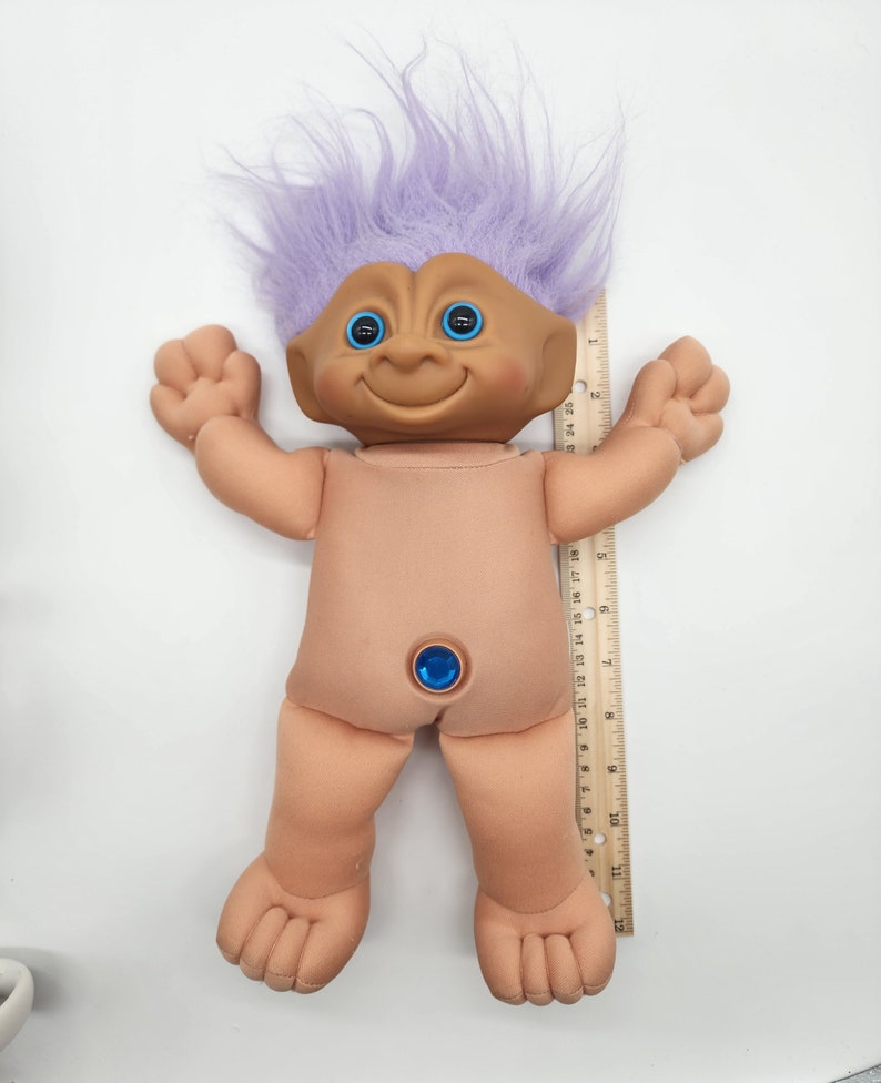 May include: A purple haired troll doll with blue eyes and a blue gem on its belly. The doll is about 12 inches tall and is standing next to a ruler.