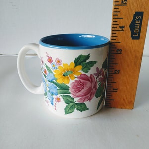 May include: White ceramic mug with a blue rim and a floral design featuring pink, yellow, and blue flowers.