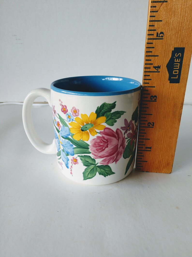 May include: White ceramic mug with a blue rim and a floral design featuring pink, yellow, and blue flowers.