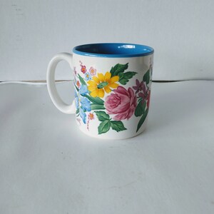May include: A white ceramic mug with a blue rim and a floral design featuring pink, yellow, blue, and green flowers.