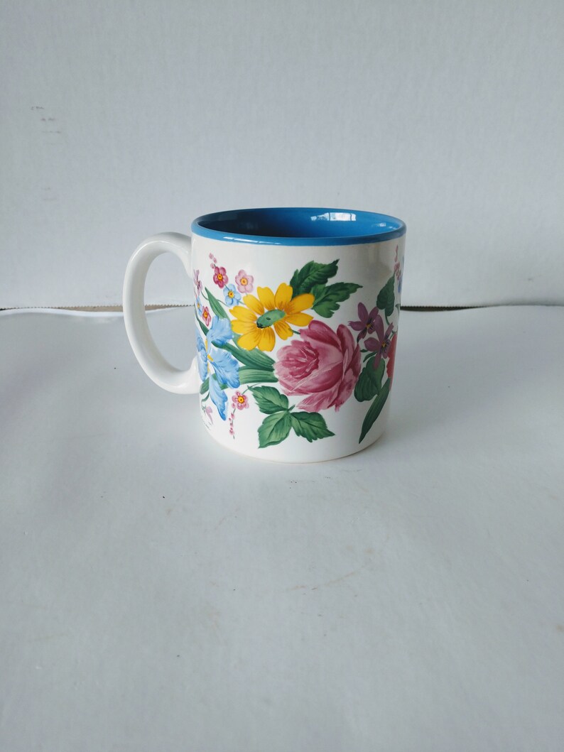 May include: A white ceramic mug with a blue rim and a floral design featuring pink, yellow, blue, and green flowers.